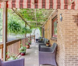 Cozy outdoor porch featuring wicker furniture, hanging plants, and a brick wall, perfect for relaxing and entertaining. Outdoor porch with wicker chairs, potted plants, and a striped awning.