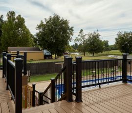 Beautiful backyard deck with black railing, overlooking a pool and lush green trees. Custom outdoor deck with black railing and pool in backyard.