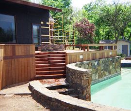 Beautiful backyard with a custom wooden deck, stone wall, and swimming pool, showcasing professional outdoor construction by Deckworks. Custom outdoor pool with wooden deck and stone wall by Deckworks.